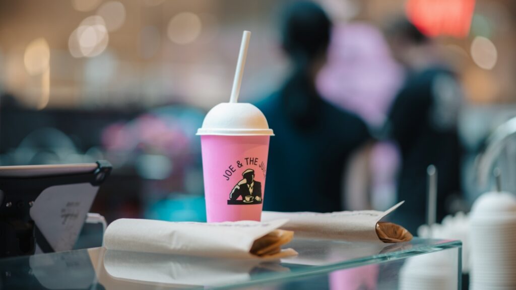 Rosa take away-mugg på cafédisk med inslagna smörgåsar och suddig bakgrundsmiljö.