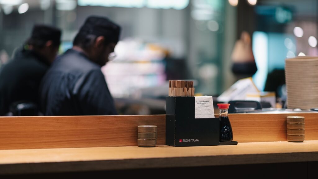 Sushirestaurang med serveringsdisk, sojaflaska och ätpinnar i fokus framför öppet kök.