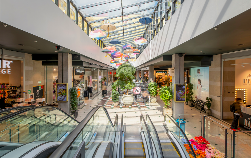 Inomhusgalleria med rulltrappor, butiker längs sidorna och färgstarka paraplyer hängande i taket under ett glastak.