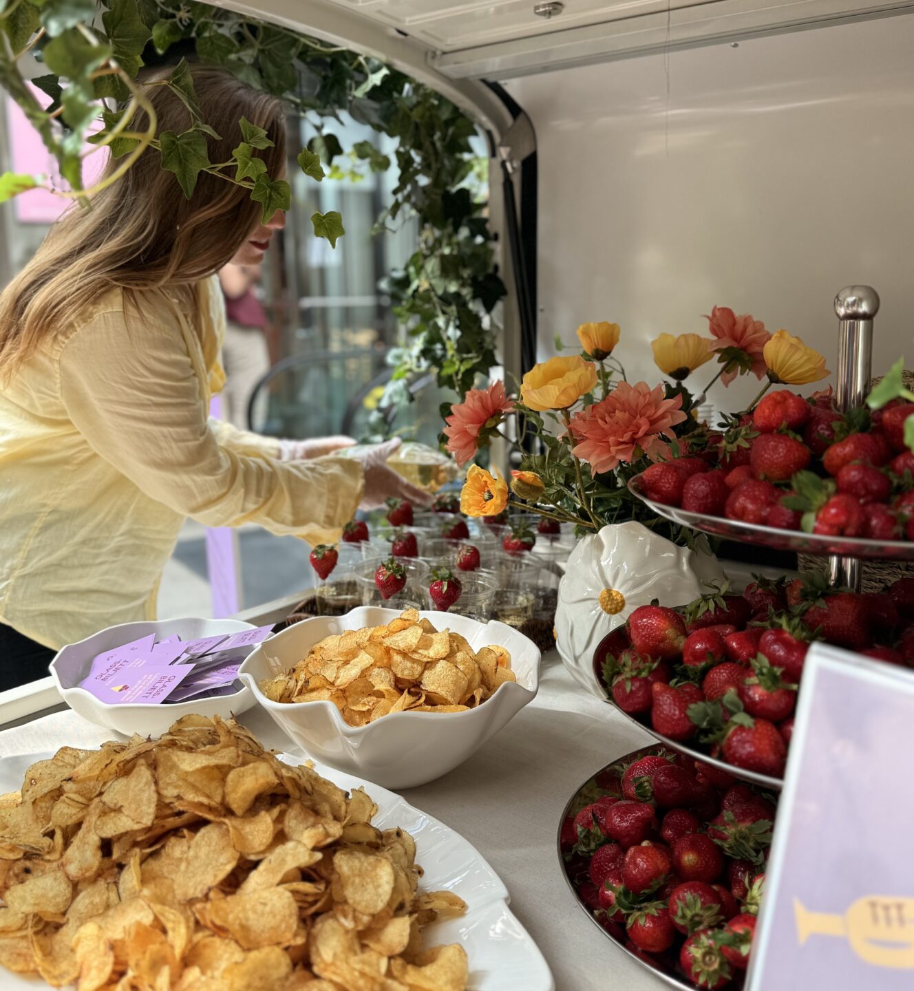 Person som plockar jordgubbar vid ett serveringsbord dekorerat med blommor och grönska, med skålar av jordgubbar, chips och drycker uppställda.